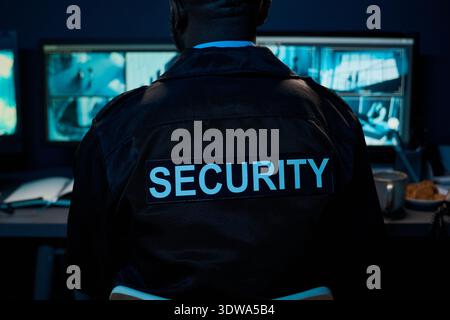 Black man middle aged sitting at security desk monitoring surveillance cameras, wearing security uniform with large SECURITY text on back, working in control room environment Stock Photo