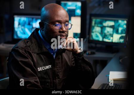 Black man wearing glasses monitoring surveillance footage on multiple computer screens in security control room, resting chin on hand while focusing on display Stock Photo
