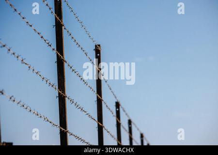 Rusty barbed wire fence with metal posts against a clear blue sky, symbolizing restriction and boundaries. Stock Photo