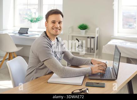 Smiling friendly doctor keyboarding on laptop computer, writing in notebook and consulting internet Stock Photo