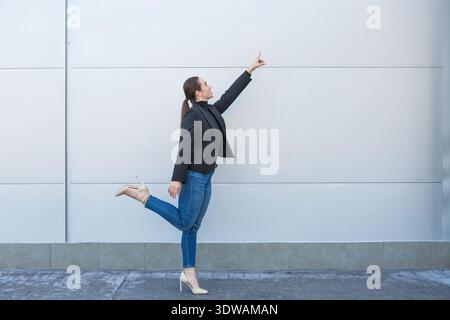 A woman is standing on one leg and pointing at a blank wall with her right hand. She is dressed in a black blazer and denim jeans. Stock Photo