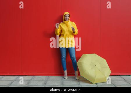 A woman stands confidently against a red wall, dressed in a yellow raincoat. She holds a zipper and has a yellow umbrella placed on the ground beside Stock Photo