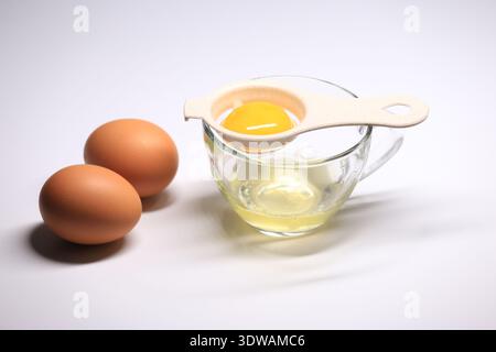 A shot of a beige egg separator holding a bright yellow yolk above egg whites in a clear glass cup, with two fresh whole eggs on a clean white surface Stock Photo