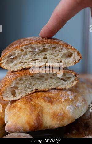 A finger gently presses the golden brown crust of a freshly baked ciabatta bread loaf. Two halves of the loaf are stacked, revealing the soft, airy interior with irregular holes. Stock Photo