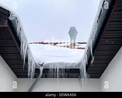 Large icicles hanging from a snow-covered residential roof and gutter during winter freeze conditions Stock Photo