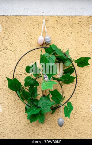 Simple Easter wreath made of green leaves and hanging pastel eggs on textured yellow wall. Spring holiday decoration and festive home design. Stock Photo