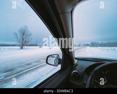 View from inside a car driving along a snowy country road next to a field and a lone tree. Winter landscape seen through the windshield and side Stock Photo