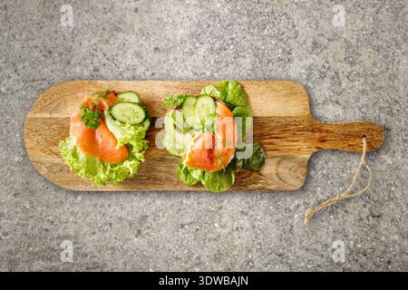 Two toasts with salted smoked salmon, lettuce salad, avocado and cucumber slices, dill and parsley on wooden board over granite stone table. Healthy f Stock Photo