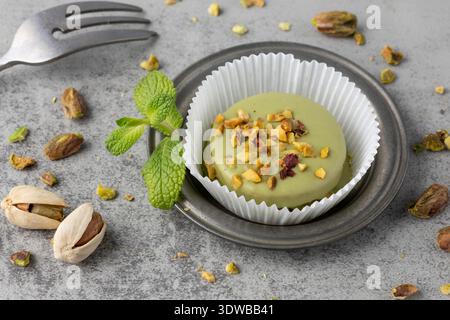 Green glazed mignon cake with crushed roasted pistachio nuts and crumbs on retro plate on grey stone background. Sweet mignon pastry  dessert. Stock Photo