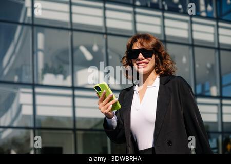 Happy businesswoman using smartphone outdoors near office building, smiling and enjoying modern professional lifestyle. Stock Photo