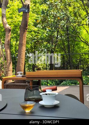 A serene tea drinking session set on a dark outdoor table with a glass teapot and ceramic cup, surrounded by lush green forest trees. Stock Photo