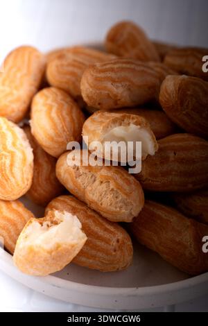 close-up of golden mini eclairs piled in a ceramic bowl: cream-filled choux pastries with ridged glossy shells and soft white filling, warm natural li Stock Photo