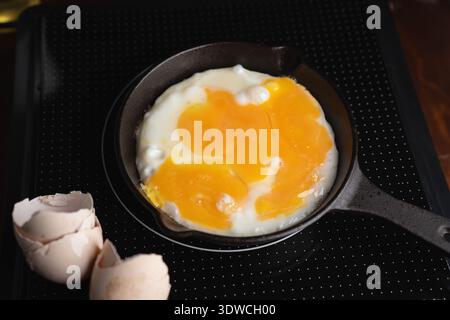 overhead close-up of a cast-iron skillet on a black dotted induction cooktop frying three eggs with glossy orange runny yolks, cracked shells nearby, Stock Photo