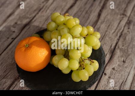 close-up still-life photograph of a plump green grape bunch beside a bright orange on a dark slate plate, resting on weathered wooden planks with warm Stock Photo