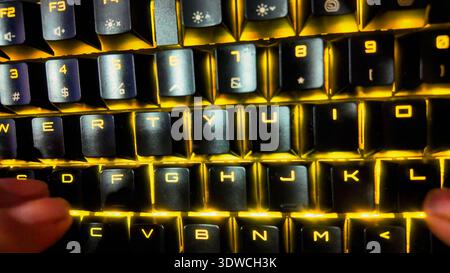 A low-angle, close-up shot of a person's hands typing on a black mechanical keyboard with dynamic, RGB backlighting in a dimly lit room. The focus is Stock Photo