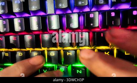 A low-angle, close-up shot of a person's hands typing on a black mechanical keyboard with dynamic, multi-color RGB backlighting in a dimly lit room. T Stock Photo