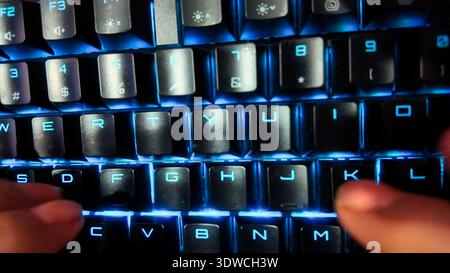 A low-angle, close-up shot of a person's hands typing on a black mechanical keyboard with dynamic, RGB backlighting in a dimly lit room. The focus is Stock Photo