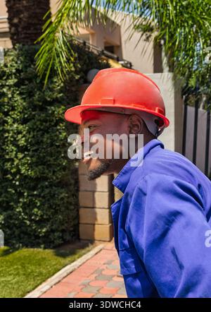 smiling happy African construction worker wearing a blue shirt and a red hard hat is captured in a low-angle, hero shot, worker close-up portrait outd Stock Photo