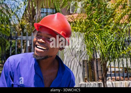 smiling Black man wearing a blue shirt and a red hard hat is captured in a low-angle, hero shot, worker close-up portrait outdoors Stock Photo