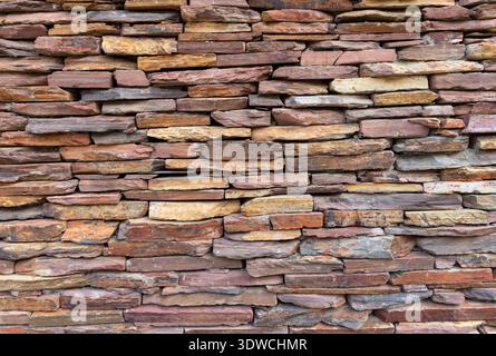dry-stacked or ledgestone wall featuring irregular, flat stones in shades of brown, tan, beige, and red, ideal for a rustic background or texture. Stock Photo