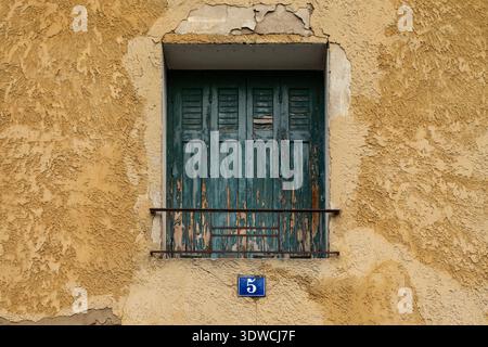 Teal wooden shutters framed by a decorative wrought iron window guard on a heavily weathered ochre wall, with a blue house number "5" plaque, showing Stock Photo