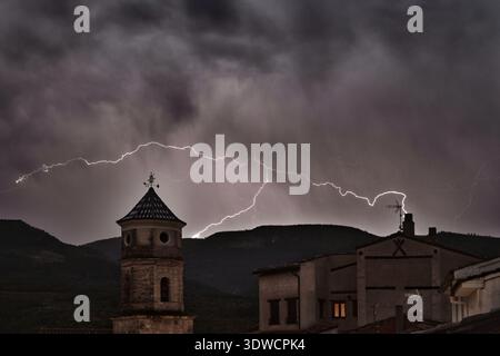 Lightning streaks across a dramatic night sky above a historic village of Casas Altas in Valencia (Spain), illuminating a bell tower and rooftops agai Stock Photo
