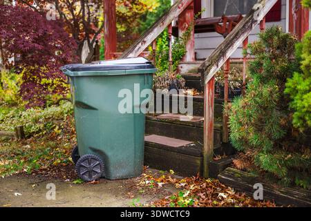 Large green garbage bin on wheels standing near the front steps of an old wooden porch surrounded by autumn foliage. Stock Photo
