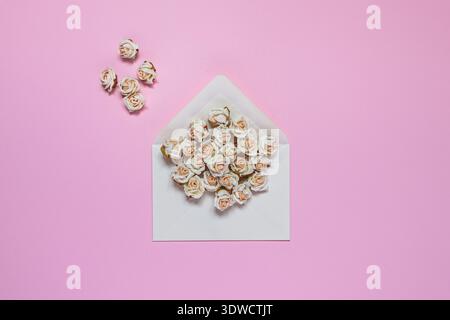 Open white envelope on a pastel pink background, overflowing with artificial beige pink roses. Additional small roses are scattered to the left of the Stock Photo