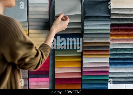 A woman wearing a brown sweater is seen from the back, reaching for a gray fabric sample among a display of multicolored velvet fabric swatches. The s Stock Photo