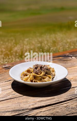 Tagliatelle with wild boar ragout is served on a white ceramic plate placed on a rustic wooden table outdoors. The setting is sunlit with a blurred ba Stock Photo