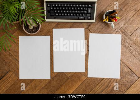 Three white A4 sheets of paper are placed on an antique parquet floor. Above them, a vintage typewriter is positioned alongside indoor plants, includi Stock Photo