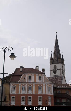In Sibiu Romania, people walk by historic buildings and a tall clock tower under a gray sky during a cloudy day Stock Photo