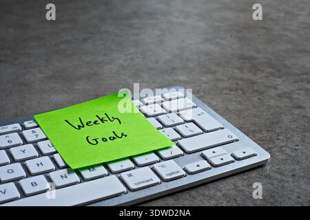 "Weekly Goals" sticky note left on keyboard. The bright note stands out against the keys, symbolising motivation, and productivity. Stock Photo