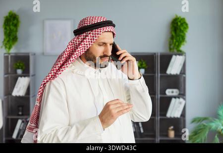 Serious Arab man in white kandura talking on smartphone, discussing important work issue in office. Stock Photo