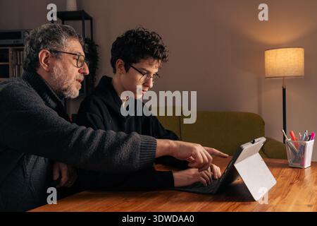 Senior father helping his teenage son study on a tablet at home. Intergenerational learning, digital education and parental guidance concept in a mode Stock Photo