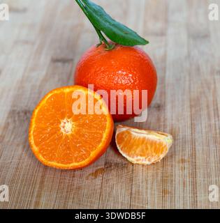 A close-up of a juicy tangerine with a green leaf, one half showing the segment structure, and a single peeled slice on a textured bamboo board. This Stock Photo