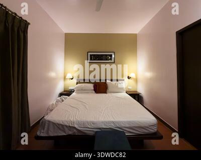 Cozy hotel room interior featuring a neatly made king bed with white sheets and pink throw pillows, flanked by bedside lamps on tan and pink walls Stock Photo