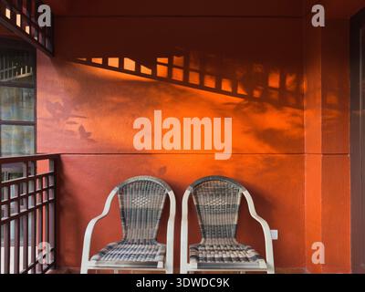 Two rattan chairs with white cushions on a textured orange-red wall corner, dramatic elongated shadows from window slats cast Stock Photo