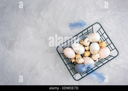 Easter eggs in metal wire basket with blue feathers on gray textured background. Top view, flat lay, copy space. Stock Photo