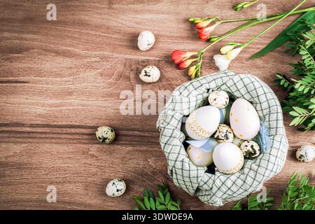Nest with painted Easter eggs, quail eggs, freesia flowers and green leaves on wooden background. Top view, flat lay, copy space. Stock Photo