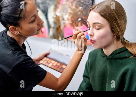 Makeup artist skillfully applies eyeshadow to a model in a studio setting. Bright colors and artistic background enhance their creative process during Stock Photo