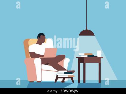 A young Black man sits comfortably in a cozy chair, focused on his laptop. Books are stacked beside him on a side table, with a cup nearby, creating a Stock Photo