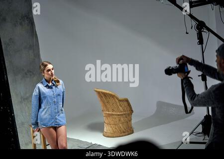A model styled in a denim shirt and shorts stands near a chair in a well-lit studio. The photographer captures the shot while focusing on the subject' Stock Photo