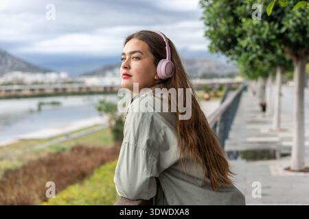 A woman with long hair enjoys a peaceful moment in the park, leaning on a railing and wearing headphones. The serene landscape provides a calming back Stock Photo