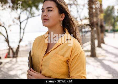 Woman enjoying remote work in a park, holding a laptop. She is surrounded by nature, emphasizing a balance between business and relaxation in an outdo Stock Photo