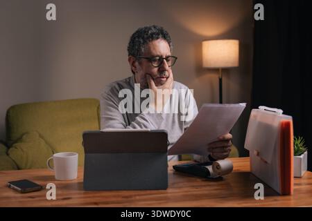 Senior man reviewing household bills and financial documents at home using tablet and calculator. Concept of budgeting, financial responsibility and m Stock Photo
