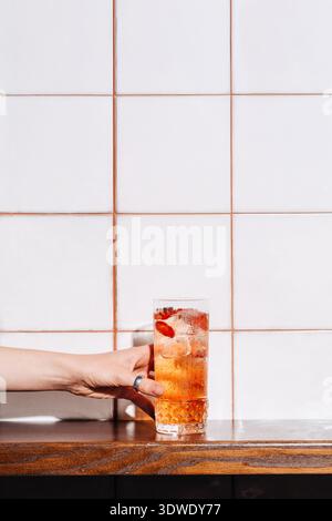 Cropped unrecognizable hand holds a strawberry cocktail garnished with ice cubes against white tiled background. The hand wears a silver ring, showcas Stock Photo