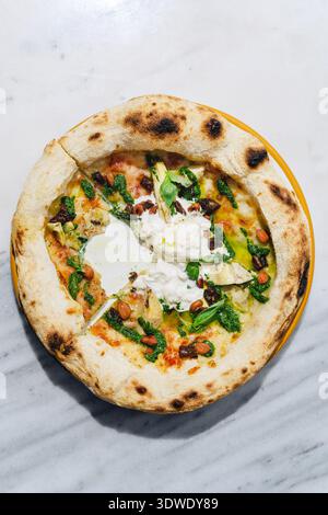 Top view of a rustic pizza topped with artichokes, almonds, pesto, and burrata cheese. The pizza is placed on a white marble table, emphasizing its fr Stock Photo