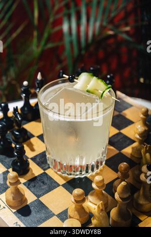 A refreshing cucumber gin fizz cocktail in a crystal glass is set on a wooden chessboard. Black and white pieces surround, with a green plant in the b Stock Photo
