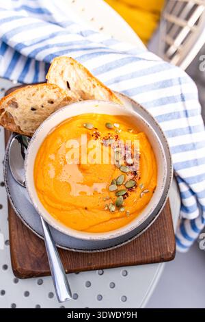 A bowl of creamy pumpkin soup garnished with seeds and served with slices of bread. The striped napkin and wooden board create a cozy, autumnal dining Stock Photo